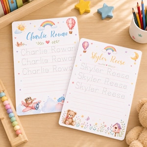 May include: Two rectangular white practice writing boards featuring colorful illustrations of rainbows, hot air balloons, and teddy bears. One board displays the name Charlie Rowan, and the other displays Skyler Reese, with dotted lines for tracing letters.