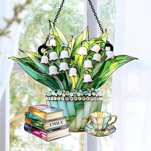 May include: Stained glass lily of the valley in a pot, hanging from a chain. The pot is light blue and green, with a decorative rim. A stack of books with the name "EMILY" is next to a teacup and saucer.