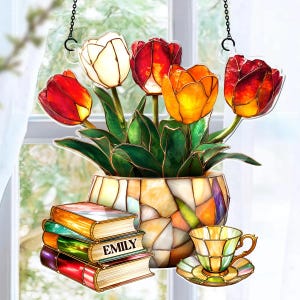 May include: A stained glass tulip window hanging with a vase of tulips, a stack of books with the name "EMILY", and a teacup and saucer. The tulips are red, orange, and white. The vase and teacup have a mosaic design.