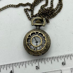 May include: Antique bronze-colored pocket watch pendant on a matching chain. The watch face is white with black numerals and hands. The watch case has an ornate, openwork design. The chain is approximately 24 inches long.