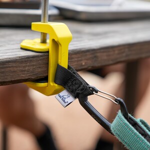 May include: A yellow table clamp with a black screw and a black and teal strap. The clamp is attached to a wooden table. The strap has a carabiner clip and a tag that says "Table Tether".