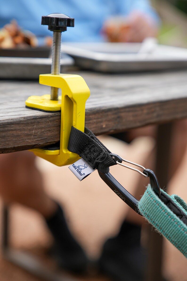 May include: A yellow table clamp with a black screw and a black and teal strap. The clamp is attached to a wooden table. The strap has a carabiner clip and a tag that says "Table Tether".