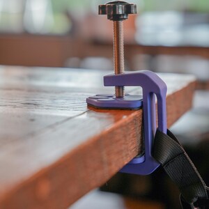 May include: A purple clamp with a black handle and threaded rod is attached to a wooden surface. The clamp is designed to secure objects, with a black strap visible on the side. The wooden surface is brown.