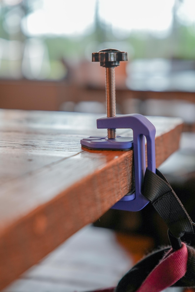May include: A purple clamp with a black handle and threaded rod is attached to a wooden surface. The clamp is designed to secure objects, with a black strap visible on the side. The wooden surface is brown.