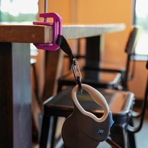 May include: A retractable dog leash with a black and white patterned leash is attached to a purple clamp. The clamp is secured to a wooden table. The leash handle is black and white with a white grip.