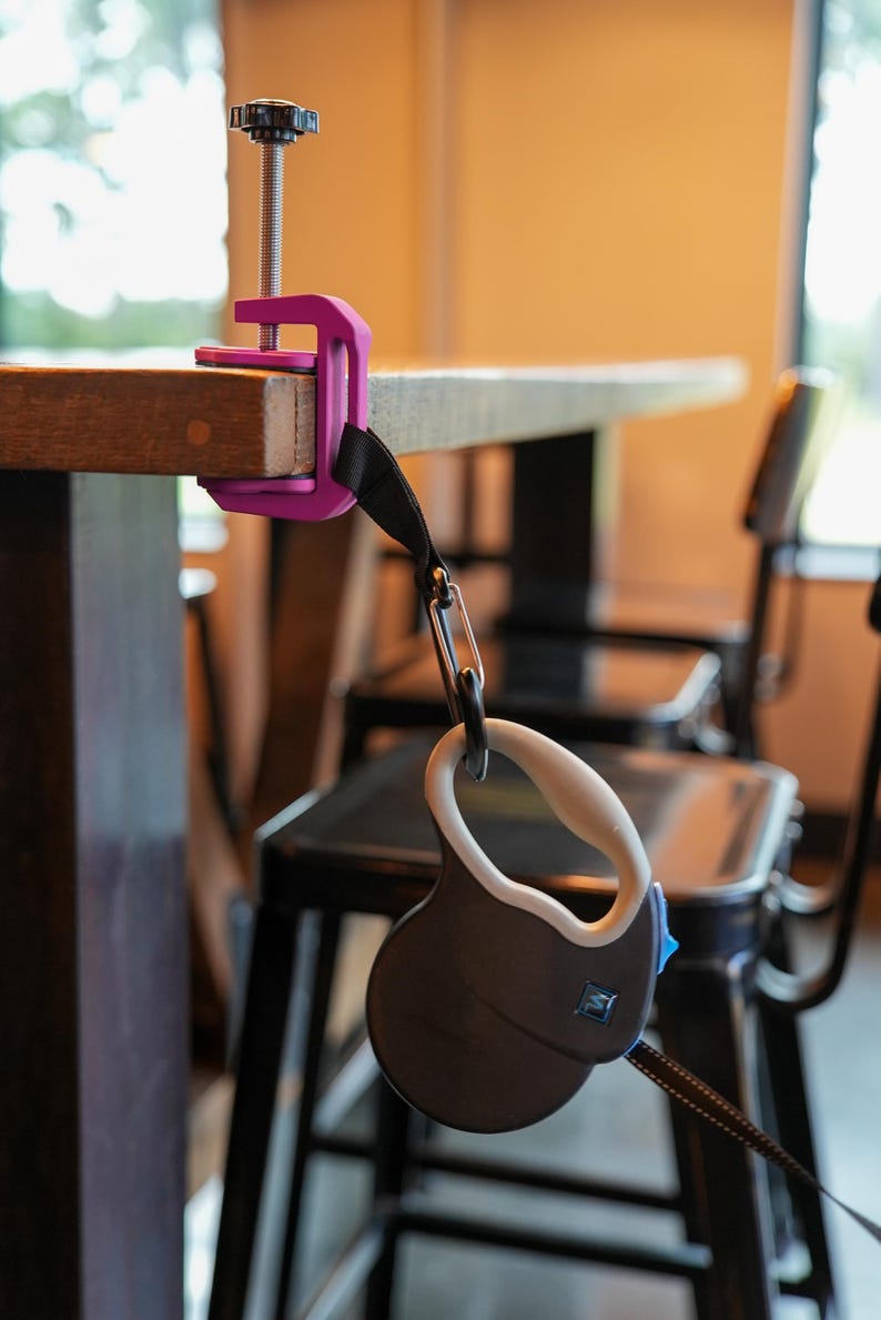 May include: A retractable dog leash with a black and white patterned leash is attached to a purple clamp. The clamp is secured to a wooden table. The leash handle is black and white with a white grip.