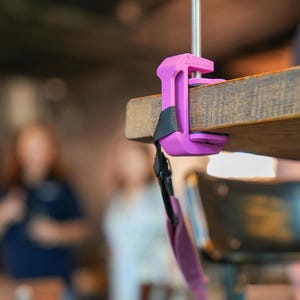 May include: A purple clamp attached to a wooden surface. The clamp has a black strap and a threaded rod with a black knob. The background is blurred, showing a cafe setting.