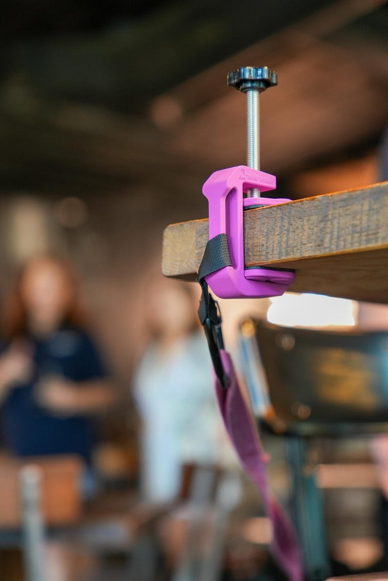 May include: A purple clamp attached to a wooden surface. The clamp has a black strap and a threaded rod with a black knob. The background is blurred, showing a cafe setting.