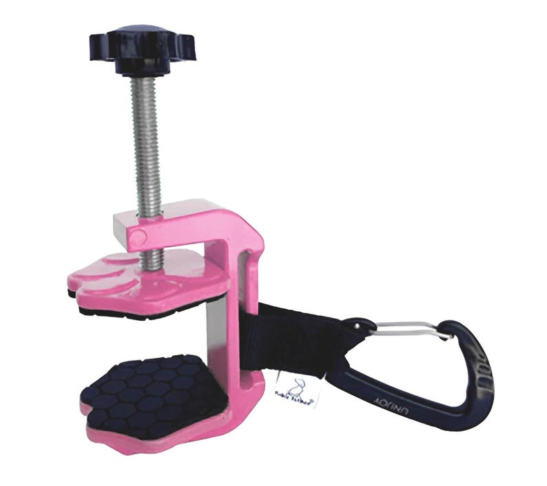 May include: A pink clamp with a black screw and handle. The clamp has a black strap and a black carabiner clip. The clamp has a black rubber pad on the bottom and a black rubber pad on the top.