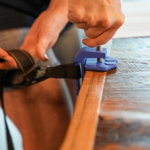 May include: A blue clamp securing a black strap to a wooden surface. The clamp has a screw mechanism for tightening. The wood is a warm brown color, and the clamp is being adjusted.