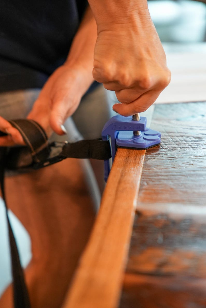 May include: A blue clamp securing a black strap to a wooden surface. The clamp has a screw mechanism for tightening. The wood is a warm brown color, and the clamp is being adjusted.