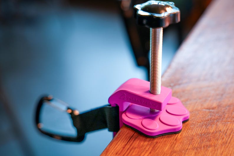 May include: A pink clamp with a black handle and a silver screw. The clamp is attached to a black strap and a wooden surface. The clamp has a paw-shaped base.