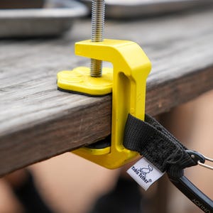 May include: A bright yellow table clamp with a black handle and screw. The clamp is attached to a wooden table. A black strap with a metal clip and a white tag that says "Table Tether" is attached to the clamp.