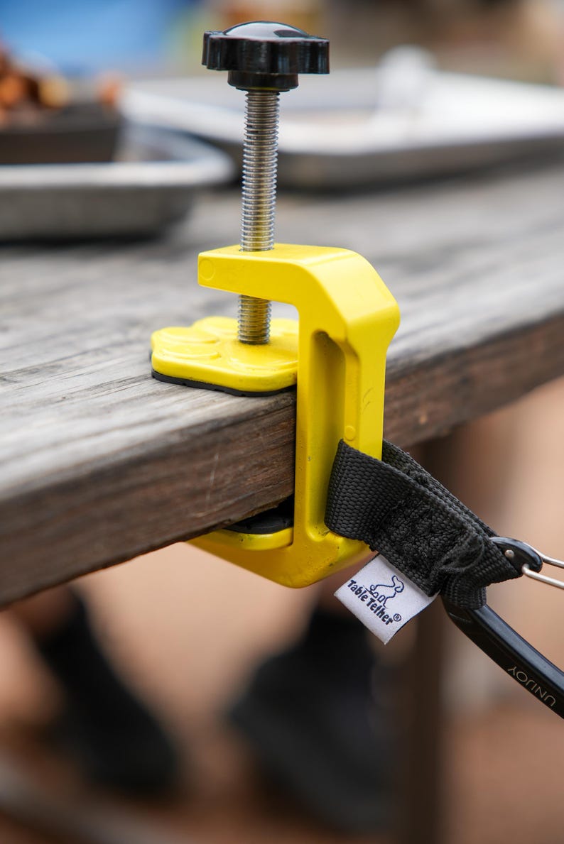 May include: A bright yellow table clamp with a black handle and screw. The clamp is attached to a wooden table. A black strap with a metal clip and a white tag that says "Table Tether" is attached to the clamp.