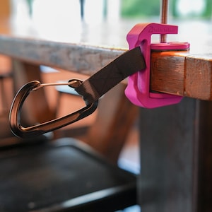 May include: A bright pink clamp-style accessory with a black strap and a carabiner clip, attached to a wooden table. The clamp has a threaded rod and a black knob for tightening. The brand name "VisibleYeller" is printed on the clamp.