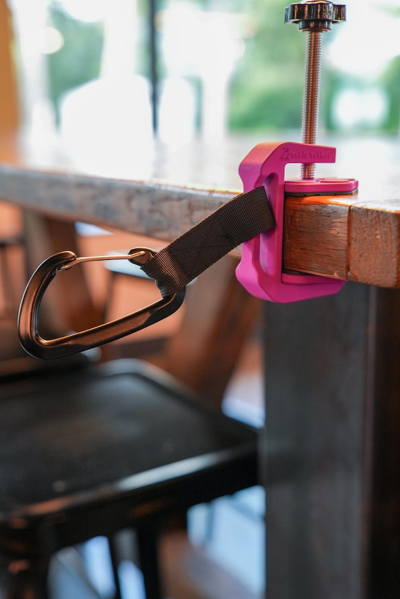 May include: A bright pink clamp-style accessory with a black strap and a carabiner clip, attached to a wooden table. The clamp has a threaded rod and a black knob for tightening. The brand name "VisibleYeller" is printed on the clamp.