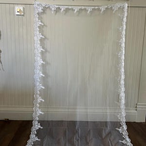 May include: A sheer white wedding veil with a delicate floral lace trim. The veil is rectangular and hangs vertically, showcasing the intricate lace design along the edges. The veil is displayed against a light-colored wall and wooden floor.
