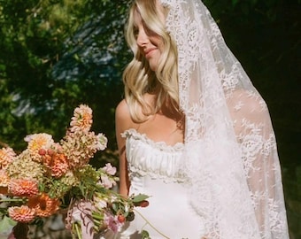 May include: A white strapless gown with a ruffled neckline and a long, intricate white lace veil draped over the head. The garment is paired with a large, colorful bouquet of orange, pink, and green flowers featuring dahlias and greenery.