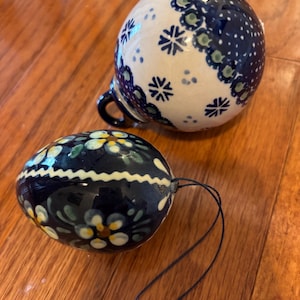 Könnte beinhalten: Ein kugelförmiges Keramikornament mit weißer Basis, blauen Schneeflockenmustern und einer dunkelblauen Schlaufe sowie ein eiförmiges Keramikornament mit dunkelblauem Hintergrund, handgemalten weißen und gelben Blumenmotiven und einer schwarzen Schnur.