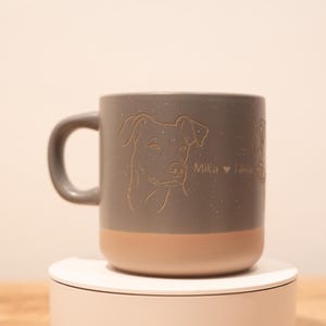 Könnte beinhalten: Ein zweifarbiger Keramikbecher mit einem gesprenkelten grauen oberen Teil und einem einfarbigen beigen Boden. Die graue Oberfläche zeigt eine fein gravierte Illustration eines Hundegesichts und den Text "Mika ♥ Toby" auf der Seite.