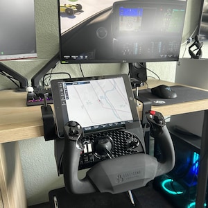 May include: A black and gray Honeycomb Aeronautical flight simulator yoke and control panel setup. The yoke is mounted on a black stand, with a tablet displaying a map. A computer monitor displays a flight simulation game.