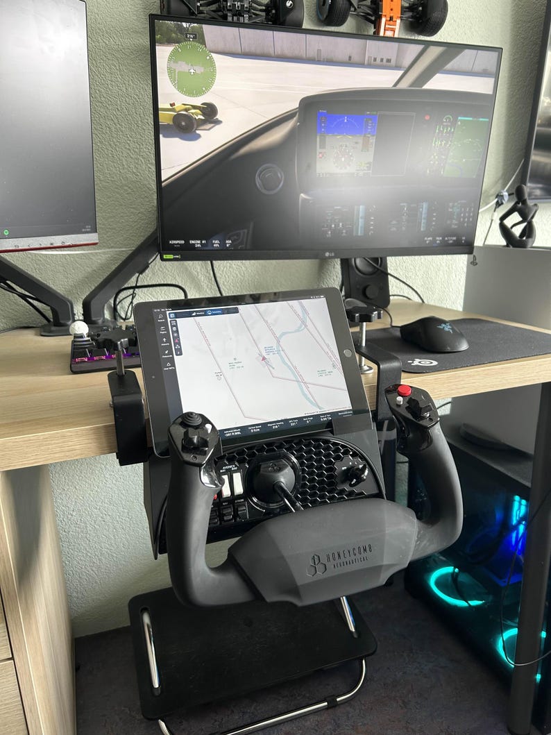 May include: A black and gray Honeycomb Aeronautical flight simulator yoke and control panel setup. The yoke is mounted on a black stand, with a tablet displaying a map. A computer monitor displays a flight simulation game.