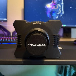 Puede incluir: Un estabilizador de cámara MOZA negro con el nombre de la marca en letras blancas en la parte delantera. El estabilizador tiene un diseño en forma de X con lados acanalados. El fondo incluye un monitor de computadora que muestra una imagen espacial.
