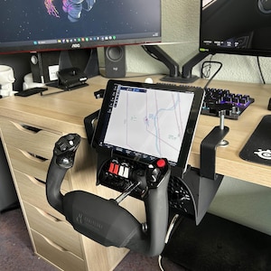 May include: Black flight simulator yoke and throttle quadrant mounted to a desk. The yoke has a black handle and the throttle quadrant has red and white switches. A tablet displaying a map is mounted above the controls.