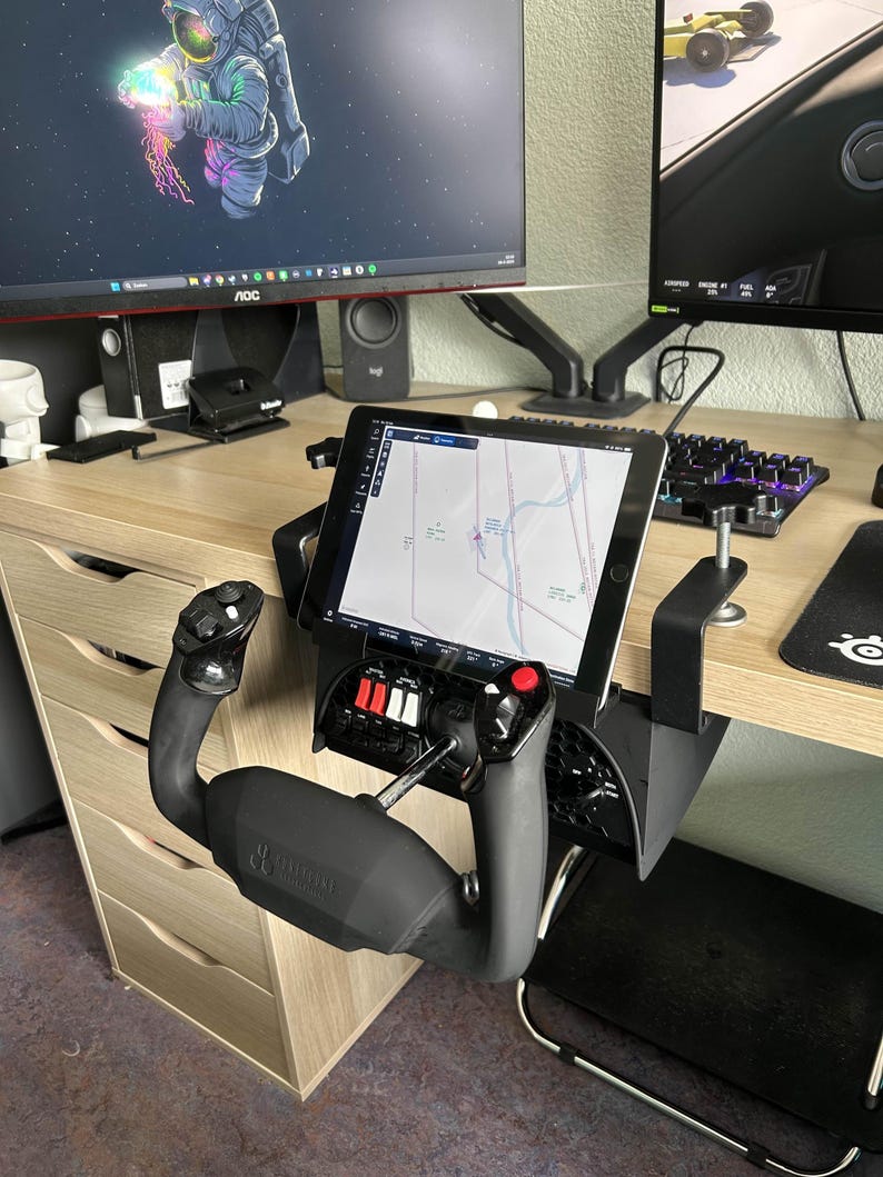 May include: Black flight simulator yoke and throttle quadrant mounted to a desk. The yoke has a black handle and the throttle quadrant has red and white switches. A tablet displaying a map is mounted above the controls.