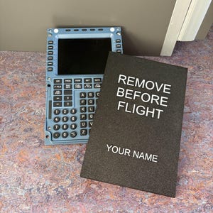 May include: A blue and black aircraft control panel with a screen, buttons, and a keyboard. A black card with the text "REMOVE BEFORE FLIGHT" and "YOUR NAME" is next to it.