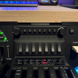 Puede incluir: Panel de control de simulador de vuelo negro con varios interruptores, perillas e indicadores iluminados. Incluye controles de engranajes, flaps, piloto automático y motor. El panel está montado debajo de una superficie de madera.