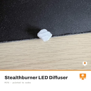 Peut inclure: Un petit diffuseur de LED Stealthburner blanc translucide avec une forme géométrique à facettes multiples. L'objet présente une surface texturée avec des lignes de couche d'impression 3D visibles, conçu pour adoucir et diffuser la lumière.