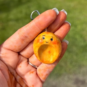 May include: A handmade, yellow-orange charm with a small face and a silver keyring. The charm has a rounded shape with a small indentation and blush-colored cheeks. The background is a blurred green.