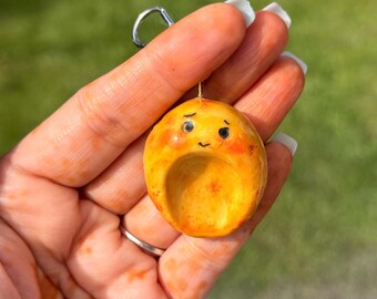 May include: A handmade, yellow-orange charm with a small face and a silver keyring. The charm has a rounded shape with a small indentation and blush-colored cheeks. The background is a blurred green.