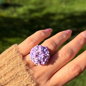 Puede incluir: Un anillo artesanal con un racimo de pequeñas cuentas en forma de flor de color lila claro. El anillo se lleva en un dedo. El fondo es un césped verde borroso.