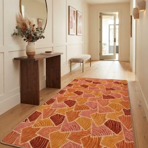 May include: A rectangular area rug featuring a repeating geometric pattern of fan-shaped segments in shades of orange, red, pink, and yellow. The rug has a textured surface with a finished border, laid flat on a light-colored wooden floor.