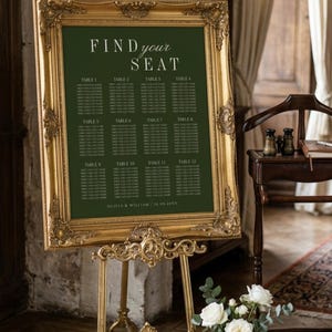 Puede incluir: Un cartel de distribución de asientos de boda verde oscuro con texto blanco que dice FIND YOUR SEAT, números de mesa del 1 al 12 y los nombres OLIVIA & WILLIAM | 18.08.20XX, en un marco dorado rectangular ornamentado sobre un caballete de metal dorado.