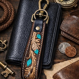 Puede incluir: Un llavero occidental de cuero hecho a mano con un diseño hecho a mano. La correa de cuero marrón presenta un patrón de hojas con detalles turquesa y está sujeta a un cierre de latón vintage. El llavero se muestra con una cartera negra y otros artículos de cuero.