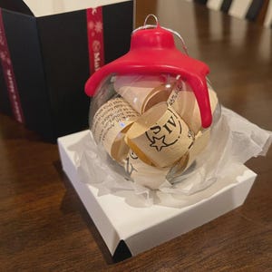 May include: A clear glass Christmas ornament with a red top, filled with rolled paper. The ornament sits on a white box with tissue paper, next to a black box with red ribbon. The rolled paper has text and a star.