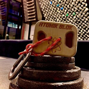 May include: A wooden Atomik Blok training tool with red rope and a carabiner, resting on a stack of weight plates. The Atomik Blok has the brand name printed on it and a graphic of an atom. The background includes a climbing wall.