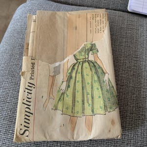 Puede incluir: Un paquete de patrones de costura Simplicity vintage. El paquete presenta una ilustración rasgada de un vestido verde con un patrón de cuadros y detalles florales. El paquete está impreso con la palabra "Simplicity" en negrita.