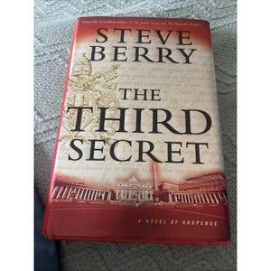 May include: A hardcover book titled The Third Secret by Steve Berry. The cover features a red and white gradient background with a gold papal emblem, black text, and a photographic illustration of St. Peter's Square at the bottom of the cover.