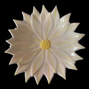 May include: White ceramic decorative bowl in the shape of a flower. The bowl has a yellow center and yellow accents on the petals. The bowl is set against a black background.