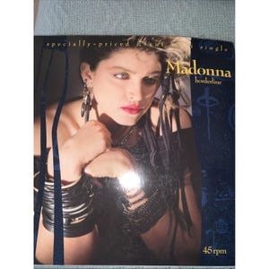 May include: A vintage Madonna "Borderline" single record cover. The cover features a portrait of Madonna with dark hair, red lipstick, and multiple bracelets. The text "Madonna borderline" is displayed in gold. The record plays at 45 rpm.