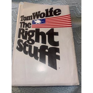 May include: A hardcover book titled The Right Stuff by Tom Wolfe with a white cover. The text is printed in large black letters. A graphic of a stylized American flag with a white eagle silhouette on a blue field is positioned above the title.