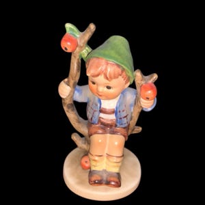 May include: A vintage porcelain figurine of a young boy in a green hat and blue jacket, holding a branch with red apples. The figurine is seated on a tree branch, with a light brown base.