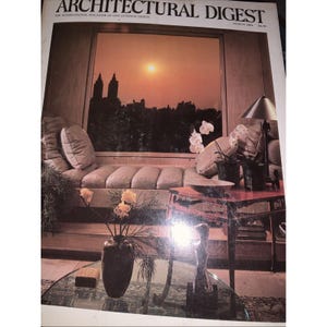 Może przedstawiać: Okładka magazynu Architectural Digest przedstawia widok zachodu słońca przez okno. Scena we wnętrzu obejmuje sofę z poduszkami, stół ze szklanym blatem i wazon z kwiatami. Widoczny jest tytuł i data wydania magazynu.