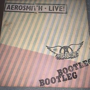 May include: Aerosmith Live! Bootleg album cover. The cover features a striped design in pastel colors, with the band's logo and the word "BOOTLEG" prominently displayed. The top section has the text "AEROSMITH LIVE!"