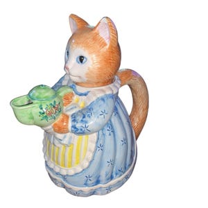 Könnte beinhalten: Eine keramische Teekanne in Form einer Katze mit orangefarbenem Fell und blauen Augen. Die Katze trägt ein blaues Kleid mit weißen Sternmustern und hält eine grüne Teekanne mit floralen Designs. Der Griff ist ein geschwungener Schwanz.