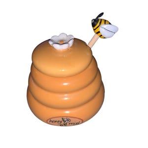 Puede incluir: Un tarro de miel de cerámica con forma de colmena, con una flor blanca en la parte superior. Un cucharón de miel de madera con una abeja de dibujos animados se inserta en el tarro. El tarro tiene las palabras "honey miel".